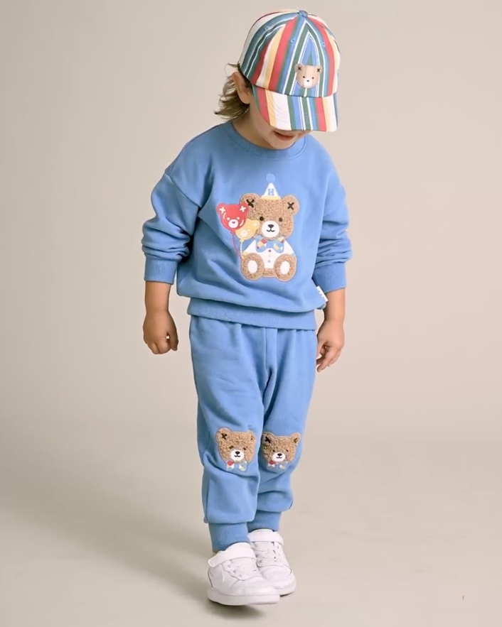 Child wearing a blue outfit with teddy bear designs and a colorful striped hat on a beige background