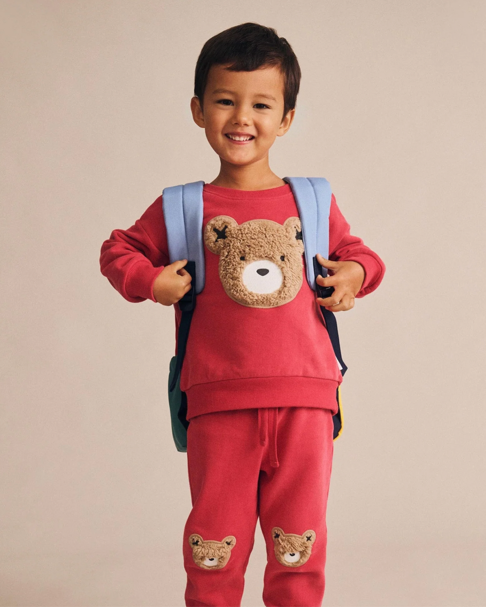 Child wearing a red outfit with bear designs and a backpack on a beige background