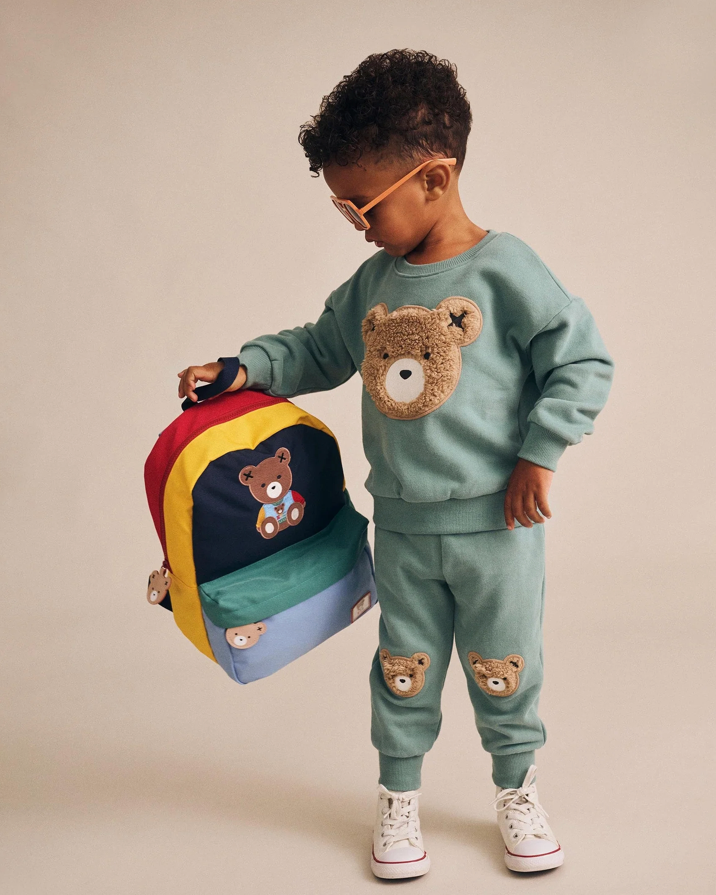 Child wearing a green sweater and pants with teddy bear patches, holding a colorful backpack.