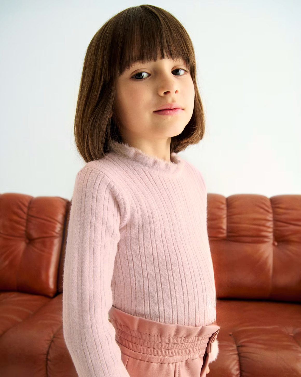 Young girl in a pink sweater standing in front of a brown couch.