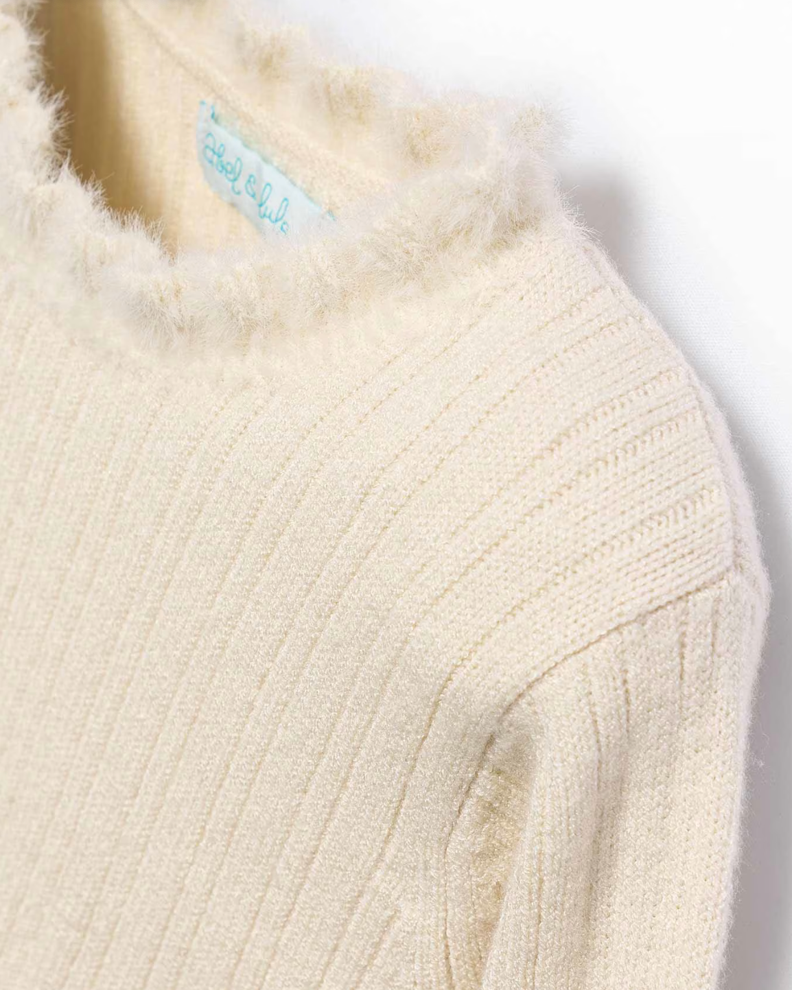 Close-up of a cream-colored ribbed knit sweater with a brand label on a white background
