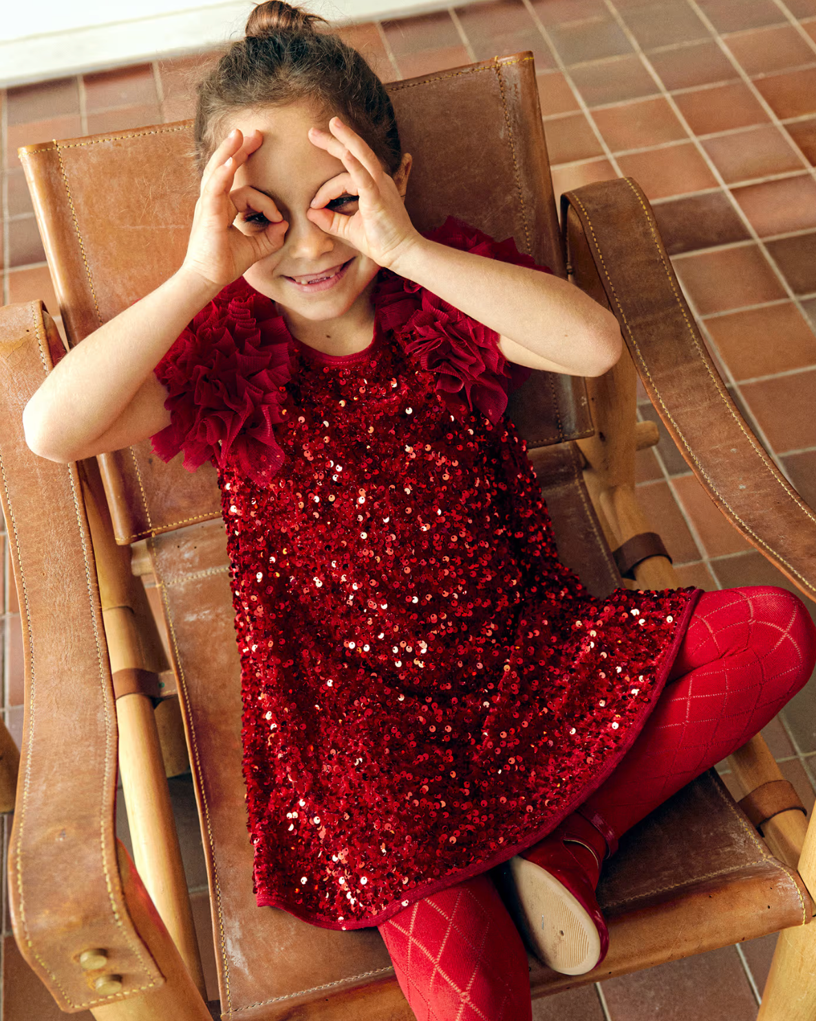 Child wearing a red sequin dress sitting on a wooden chair.