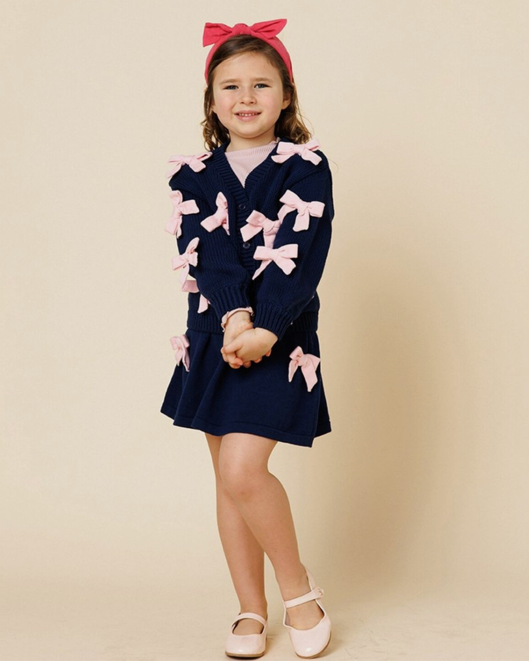 Young girl wearing a navy blue dress with pink bows against a beige background