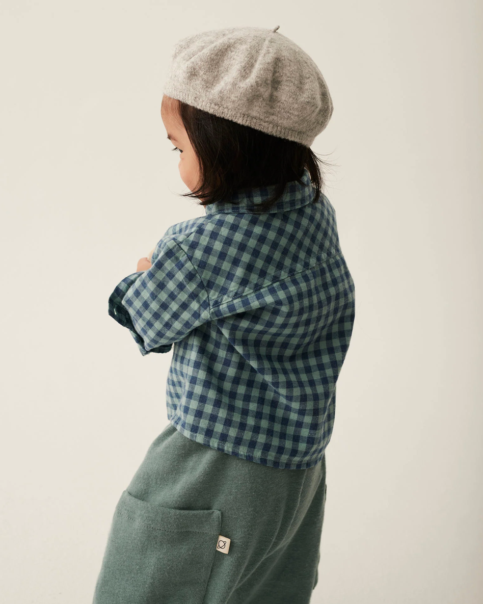Child wearing a gray beret and blue plaid shirt with green pants on a plain background