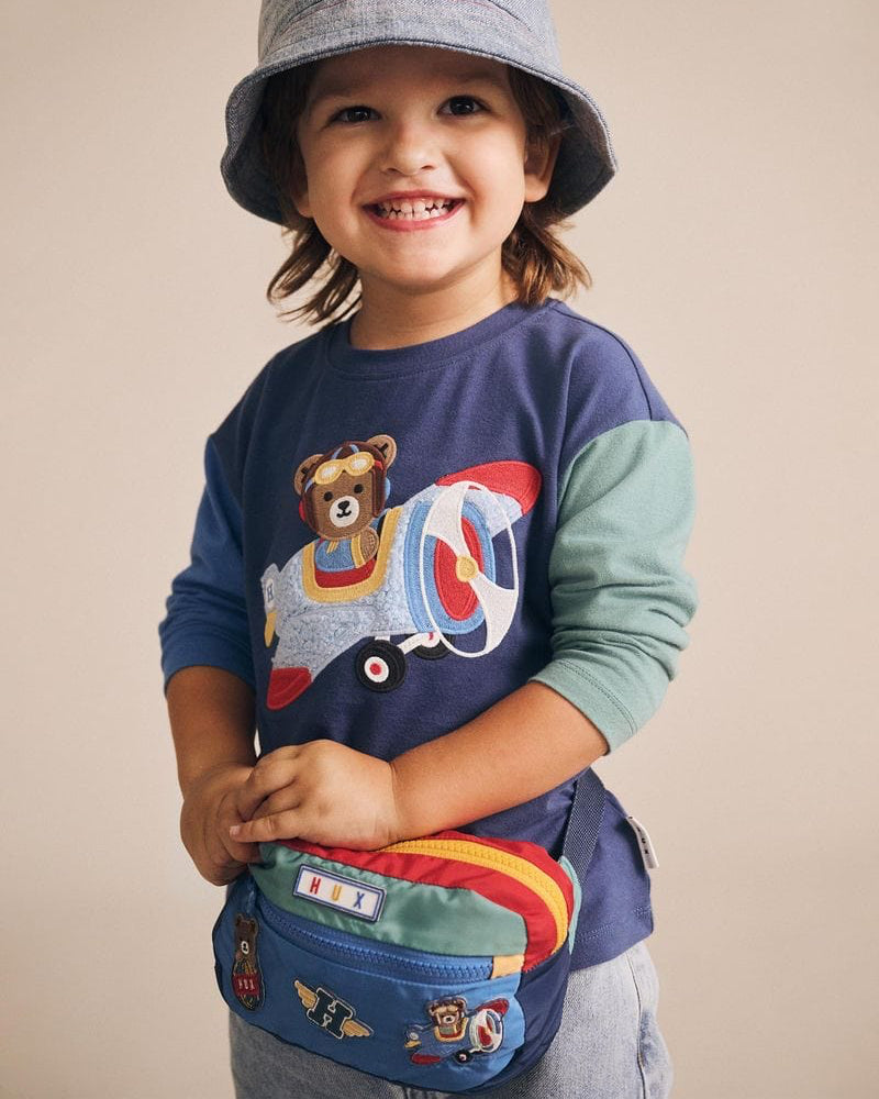 Child wearing a blue shirt with a bear design and a colorful bag, standing against a beige background.